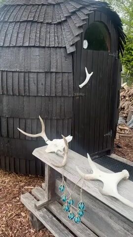 The scene shows a small, dark, barrel-shaped structure resembling a sauna or outhouse, possibly in a backyard or rural setting. The structure is covered in overlapping wooden shingles and has a rounded roof. A small arched window is visible near the top of the door. Several deer antlers decorate the area. One is mounted on the door, while two others lie on a wooden bench in front of the structure. A string of small, blue, glass balls hangs from the bench. The ground is covered with wood chips and a pile of cut firewood is visible in the background. The scene appears calm and still, with no discernible movement or people present. The lighting is natural, and it appears to be a slightly overcast day.