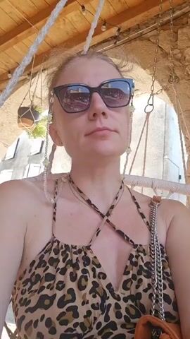 A woman wearing sunglasses and a leopard-print dress is sitting in a swing chair outdoors. The swing chair has a wooden ceiling with lights above. The woman is looking up and to her right. She has a beaded chain attached to her glasses. A purse with a chain strap is visible to her right.