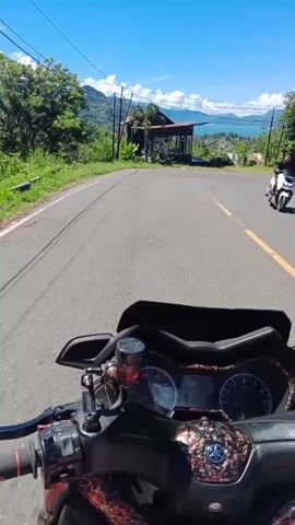 The video is shot from the perspective of a person riding a motorcycle. In the foreground, the handlebars and dashboard of the motorcycle are visible. The road ahead is paved and curves to the right. On the right side of the road, there is a small, rustic structure with a tin roof, surrounded by greenery. Beyond this structure, a body of water, likely a lake or sea, is visible, with mountains in the distance. The sky is clear and blue, with a few wispy clouds. The overall scene suggests a scenic ride through a rural or mountainous area. There are no people visible in the video, only the motorcycle and the natural environment with some man-made structures.