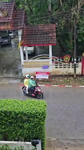 The video shows a person driving a motorcycle on a wet asphalt road. Another person is riding as a passenger behind the driver. Both individuals are wearing helmets and raincoats. A building with a red-tiled roof and a white wall with decorative elements is visible on the left side of the road. Trees and lush green foliage are present in the background. The road appears to be in a somewhat rural or suburban area, with power lines overhead and scattered debris on the ground, possibly from fallen branches or leaves. Another motorcycle is parked on the right side of the road. The lighting suggests it may be daytime, possibly overcast due to the rain.
