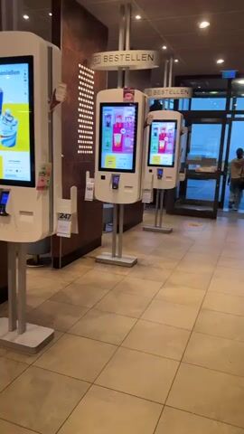 The video shows the interior of a McDonald's restaurant. In the foreground, there are several self-order kiosks with touchscreens displaying menu items. The kiosks are white and have metal stands. To the left of the kiosks, a dark brown wall with a textured pattern is visible. The floor is tiled with large, beige tiles. In the background, there are tables and chairs, some of which are high-top tables with stools. The overall lighting appears to be artificial, with some yellow ceiling lights visible. The environment appears clean and modern, consistent with a fast-food restaurant. There are no people clearly visible in the immediate foreground, but there appear to be a few customers seated at tables in the background, though their features are not discernible due to the distance and blurriness of the footage. The video pans from the kiosks to the seating area, offering a glimpse of the restaurant's layout and ambiance.