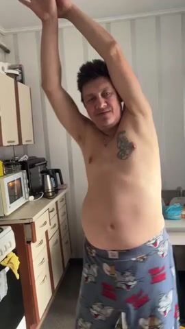 A topless man with short dark hair and tattoos on his chest and upper arm is standing in a room, stretching his arms above his head. He is wearing pajama bottoms with a cartoon pattern. The room appears to be a kitchen, with cabinets and appliances visible in the background. The man is facing the camera initially, then turns to his side and then to his back.