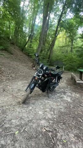 A black motorcycle is parked on a dirt path in a forest. A green picnic table with two benches is located to the right of the motorcycle. Two helmets, one red and one white, are on the table. The trees in the background are tall and lush, with green leaves. The sun is shining through the trees, creating dappled light on the ground.
