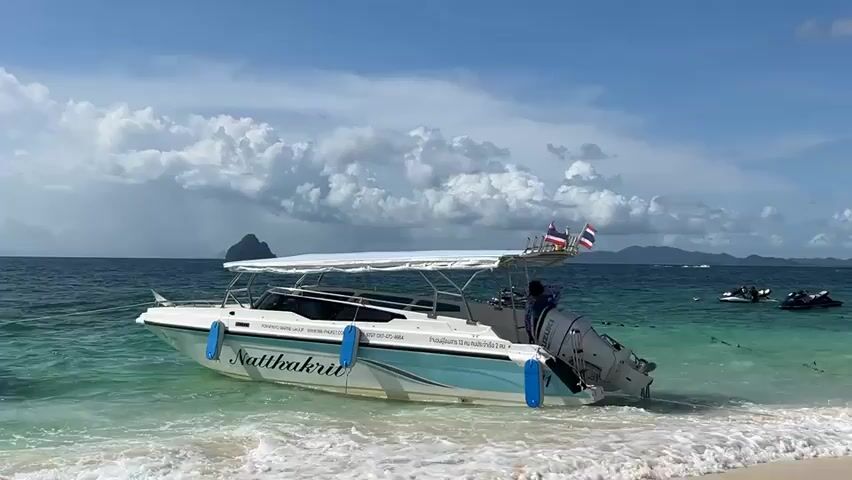 A speed boat named NATTHAKRIT is moored near a sandy beach. The boat has a white canopy supported by a metal frame, and it is partially in the shallow turquoise water. Two blue fenders hang from the side of the boat. A person in a blue shirt and dark pants is on the stern of the boat, near the outboard motor. There are two Thai flags mounted on a pole at the stern. In the background, there are other boats, including jet skis, and a hazy island in the distance. The sky is blue with scattered white clouds. Waves are gently breaking on the shore.