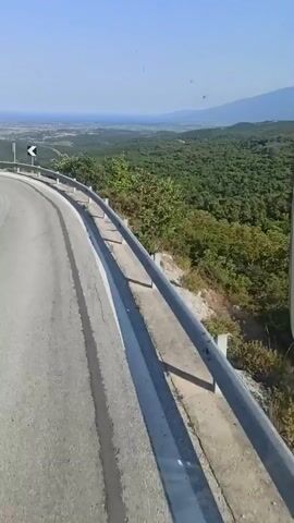 This media file depicts a scenic view from a moving vehicle on a winding road. The road curves to the left, with a guardrail running along its edge. Beyond the guardrail, a dense green forest stretches out, eventually meeting a vast expanse of blue sea in the distance. A small town or village is visible along the coast. The sky is clear and blue, suggesting a bright, sunny day. The time of day appears to be midday or early afternoon. The overall mood is one of serene travel and appreciation of natural beauty. The location is identified as Palaios Panteleimonas, Greece, which aligns with the coastal and mountainous scenery. There are no people clearly visible in the footage, and the focus is on the landscape and the journey. One visible object on the roadside is a traffic sign with a black chevron on a white background, indicating a curve ahead.
