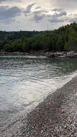 The video shows a close-up view of a pebbly beach with gentle waves lapping at the shore. The water is clear and reflects the overcast sky, which is filled with dramatic clouds. In the background, a lush green hillside covered in pine trees slopes down to the water. The scene is serene and natural, evoking a sense of peace and tranquility.