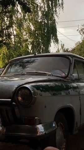 The video shows a close-up of an old, faded white and green car parked outdoors. The car has a prominent chrome grille and a circular headlight visible on the left side. The paint on the car is peeling and weathered. Trees with green foliage are visible in the background, and the sky is overcast. The camera is positioned low, focusing on the front of the car. The license plate is partially visible, showing the characters "E1340H 196 RUS". There are water droplets on the windshield. The scene appears to be shot in daylight, with soft, diffused lighting.