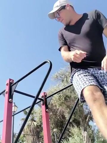A man wearing a black t-shirt, plaid shorts, a baseball cap, and glasses is performing exercises on parallel bars outdoors. The sky is clear and blue, with some trees and buildings visible in the background. The man is holding onto the bars with his hands and is lifting his legs off the ground, demonstrating strength and balance. He moves his legs up and down in a controlled manner while maintaining his grip on the bars.