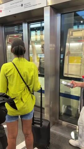 The video captures a scene at the Zurich Airport's Skymetro, where two individuals stand in front of train doors waiting to board. On the left, a woman is seen from behind, wearing a bright yellow shirt and denim shorts. She has a dark-colored crossbody bag and a black rolling suitcase. To her right stands a young man who holds a smartphone in his hand. Both individuals are facing the train doors, which reveal the interior of the train and other passengers inside. Above the doorway, a sign reads "Skymetro Flughafen Zurich Gates A,B,D. Ausgang Exit. Gepäckausgabe Baggage Claim." A green exit sign is also visible above the doorway. The setting appears to be a clean, modern airport terminal.