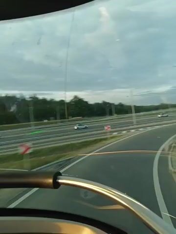 The video shows a view from inside a vehicle looking out through the windshield. The vehicle appears to be on a highway with multiple lanes. Several cars are visible on the highway in the distance. The sky is overcast with clouds. There are some lines and marks on the windshield, suggesting it may be dirty or scratched. The foreground shows a portion of the vehicle's interior, including a metal bar and some black plastic trim, likely part of the dashboard or a grab handle. The overall impression is of travel on a highway during a cloudy day.