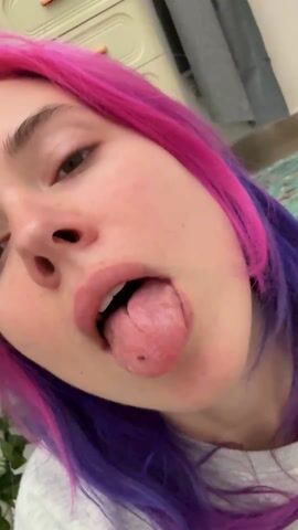 A woman with pink and purple hair sticks her tongue out at the camera. She is wearing a gray shirt. She then closes her mouth, smiles, and opens her eyes. Behind her is a beige plastic dresser with drawers and a gray curtain. The floor has a carpet with a green pattern.