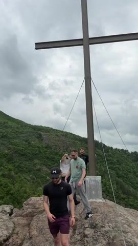 A video shows a group of four men standing near a tall metal cross on a rocky hilltop. The cross is supported by guide wires. One man is walking forward, wearing a black shirt, black baseball cap, and dark shorts. The others stand near the cross. The camera pans to reveal a panoramic view of a green valley, a town with red-tiled roofs, and mountains in the distance under a cloudy sky. It then pans down to a small shelter with a red roof on a lower level of the rocky terrain, where two people are standing. The landscape is green with vegetation and rocky outcrops.