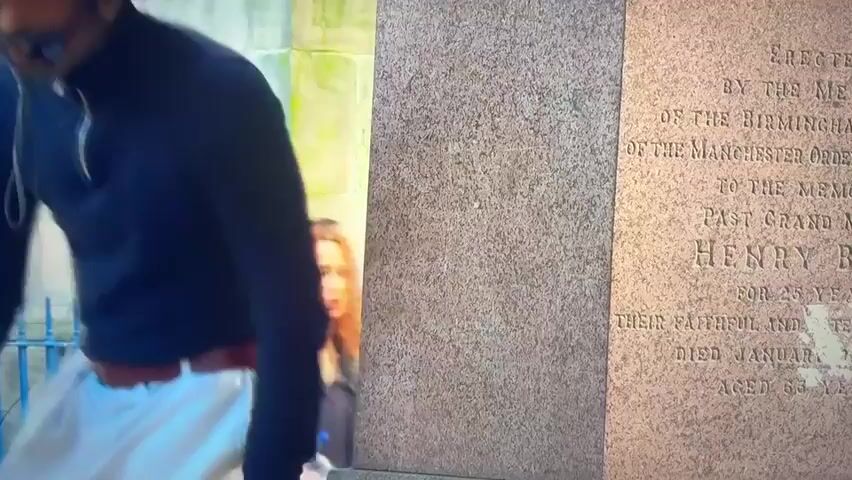 The video shows a man wearing a black turtleneck shirt, white cargo pants, and a brown belt standing next to a large stone monument. The monument has inscriptions on it, which are partially visible and appear to be in English. The man is holding something up to his face, possibly a camera or sunglasses. The background shows a building with stone architecture, a fence, and a banner that says "WELCOME TO BIRMINGHAM CATHEDRAL." The scene appears to be an outdoor urban environment.