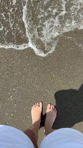 The video shows a first-person view of a person standing on a sandy beach with the ocean waves approaching their feet. The person is wearing white shorts and has bare feet. The sand is dark and wet from the water. The waves are small and foamy, creating a light white line as they recede. The sun casts a shadow to the right of the person's feet. The video is static, with no discernible movement of the person or the environment.
