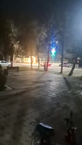 The video depicts a street scene at night, with a park on one side and a road on the other. The road has several cars passing by, and a pedestrian is walking on the sidewalk.  There is a decorative, brightly lit figure on a pole next to the sidewalk. In the park, there is a circular structure engulfed in flames. There are benches in the park and trees lining the street. In the foreground, a parked scooter is visible. The ground is covered with cobblestones. The video is taken from a slightly elevated position. The lighting is dim, with artificial light sources illuminating the scene.