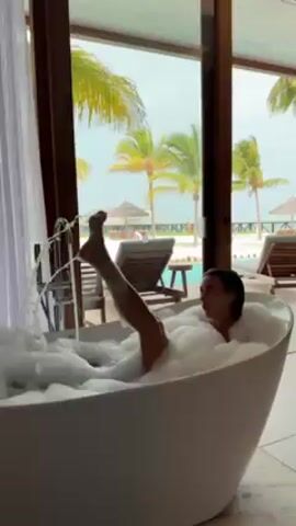 A woman is in a white oval bathtub full of bubbles. She is smiling and speaking to the camera. One of her legs is briefly raised in the air. Through a large window, a resort can be seen, including palm trees, lounge chairs, a swimming pool, and umbrellas on the beach.