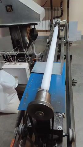 The video shows a long white cylindrical object being processed on a machine. The object is held horizontally by two rotating cylindrical supports, one at each end, which are part of the machine. The machine itself appears to be industrial, with metal components, gears, and a conveyor belt system visible in the lower left corner. To the left of the machine, there is a large white bag, possibly containing raw materials. In the background, a radiator and some electrical components are visible. The overall environment appears to be a workshop or factory setting. The white object is rotating, and its surface seems to be smooth. The video is shot from a medium distance, focusing on the processing of the object. There are no people visible in the video.