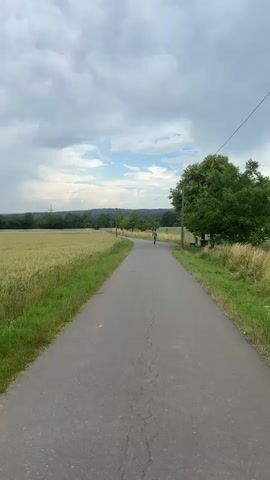 A person is seen riding a skateboard down a paved road in a rural area. The road is flanked on the left by a field of golden wheat with a few red poppies. On the right side of the road, there is grass, a large tree, and power lines. The sky is cloudy with some patches of blue sky visible. The camera is positioned behind the skateboarder, looking forward down the road.