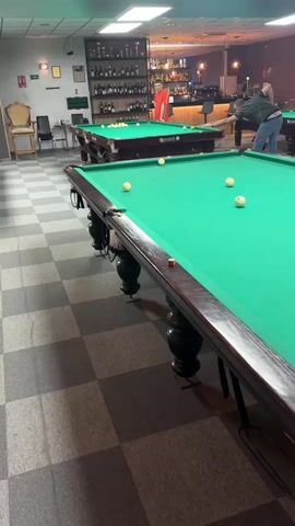 The video shows an indoor billiards room. Several billiards tables with green felt are visible on a black and white checkered floor. On one table, a man in a black shirt leans over the table, cue in hand, and prepares to make a shot at an arrangement of billiard balls. Another man in a white tank top stands to the right, watching the play. Behind them, a bar area is visible with a selection of bottles on display. Further back, two more men are playing at other tables, one in an orange shirt and another in jeans and a striped shirt. A television hangs on the wall and various trophies and decorations are spread across the room.