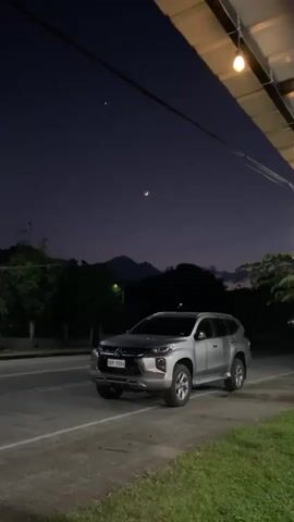A silver SUV is parked on the side of a road at night. The sky is dark blue with a crescent moon and a star visible. Trees and mountains are silhouetted in the background. A roof with lights extends over the right side of the frame.