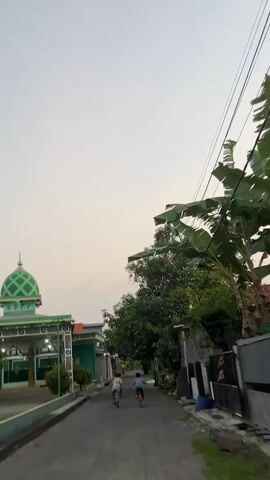 Two children are cycling down a narrow asphalt road in a village. The child in front is wearing a blue shirt and riding a red bicycle. The child behind them is wearing a light brown shirt and riding a dark blue bicycle. On the left side of the road is a building with a green dome and a white minaret, which appears to be a mosque. The building has white pillars and a covered walkway. There are also smaller green and white buildings visible. On the right side of the road, there are houses with dark fences, some with corrugated metal roofs. Large banana leaves are visible in the foreground on the right, along with power lines strung overhead. The sky is a pale, overcast gray, suggesting either early morning or late afternoon. The overall atmosphere is peaceful and typical of a rural Indonesian village scene.