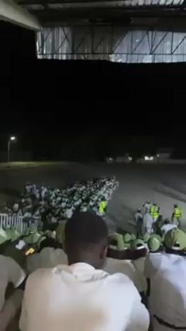 The video shows a large group of people gathered at night, possibly in a stadium or large covered area. The majority are wearing white shirts and green caps, suggesting a uniform or common affiliation. Some individuals are also wearing fluorescent yellow vests. The crowd seems to be observing something happening on the field in front of them. There are multiple rows of seated individuals, with the camera positioned among them. The setting appears to be a well-lit open space, possibly a parade ground or sports field, with some buildings visible in the background.