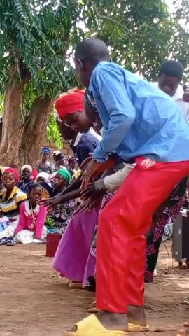 The video shows a group of people gathered outdoors, likely celebrating. Several people are dancing, some wearing traditional clothing, while others are singing and clapping.  A man with a microphone appears to be leading the singing. Many people are sitting and watching the performance. The setting seems to be a rural area, with trees and a brick building in the background.