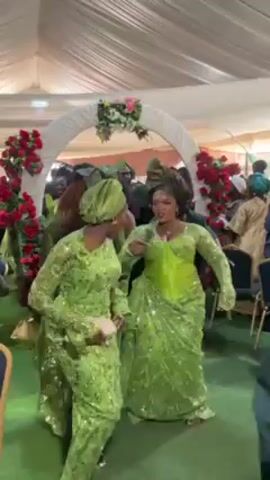 Several women in matching green, floor-length dresses with traditional headwraps dance closely together in a celebratory setting. They are under a white floral arch in what looks like a tent or large room with other partygoers in the background. The women’s outfits are detailed with patterned fabric. The dancers seem to be having a joyful time, moving to music playing in the background. The scene is brightly lit, indicating it is a festive occasion, possibly a wedding.