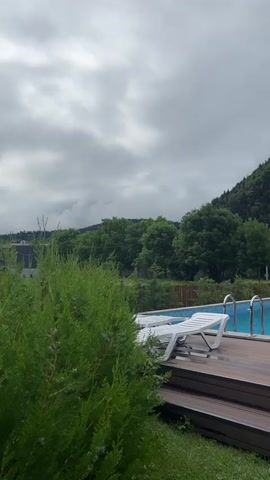 A vertical video pans across an outdoor swimming pool and a wooden deck, with a triangular wooden cabin and a forested mountain in the background. The sky is overcast and cloudy. The pool is filled with blue water and has two white lounge chairs on the deck. The deck is made of brown composite material and has steps leading down to a grassy area. A path runs along the side of the deck, leading to the back of the property. The cabin is made of logs and has a black roof. An air conditioning unit is visible on the side of the cabin. The mountain is covered in dense green trees, with clouds partially obscuring the summit.