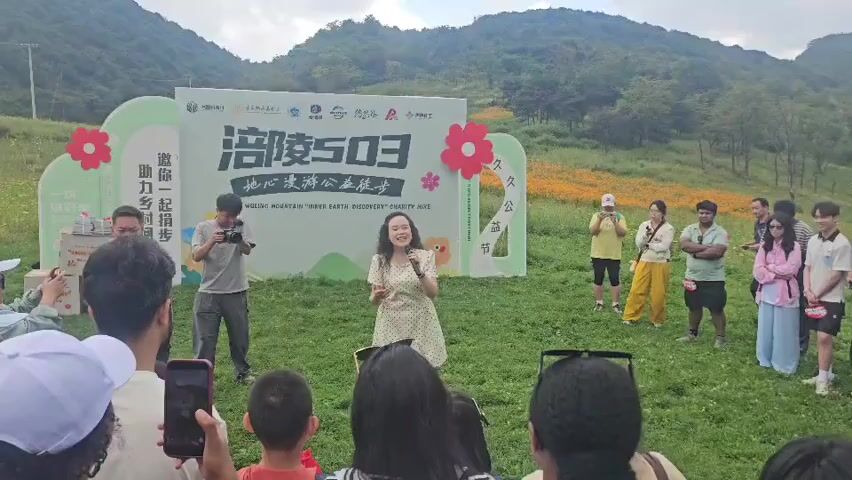 A woman in a polka-dot dress stands center frame, holding a microphone and speaking into it. She has long, dark, curly hair and is looking towards her right. She gestures with her left hand. Behind her is a large, light-colored banner with Chinese characters and some floral and mountain graphics. To the left of the woman, a man in a gray t-shirt and dark pants is filming her with a video camera. To the right of the woman, a group of people are standing, watching. Some of them are holding phones up, presumably to take pictures or videos. The woman is on a grassy area, and in the background, there are hills covered in trees and some yellow flowers. Several people are visible in the foreground, facing away from the camera and towards the woman. A few children are also among the spectators. The overall atmosphere appears to be an outdoor event.