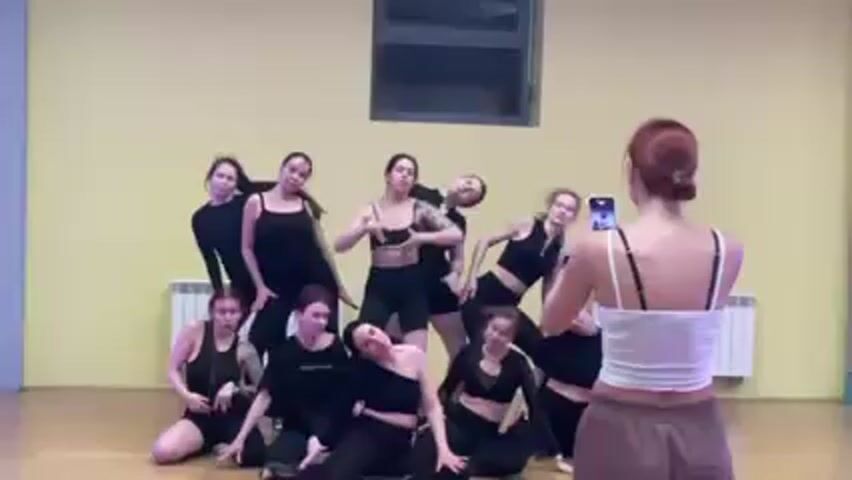 In a brightly lit, modern dance studio with yellow walls, a group of approximately ten young women dressed in various black dance outfits pose for a photo being taken by another young woman. The woman taking the photo stands facing the group, holding up her smartphone to capture the image. The dancers are arranged in a tiered formation, some standing, some kneeling, and others reclining on the floor. They are posed in dynamic, coordinated poses, displaying elements of dance technique. The studio includes features like a window with an obstructed view and a radiator along the wall. The overall setting is professional and likely related to a dance class or performance preparation.