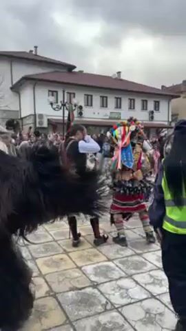 The video shows a group of people in costumes dancing in a town square. They are wearing various masks and animal furs, some with bells attached to their clothing. Most of the costumes are black or white, with occasional colorful decorations. The group is tightly packed, and they are moving energetically with synchronized steps. Behind the dancers is a crowd of onlookers, and a two-story building. The weather appears to be overcast. The surface underfoot is paved with concrete.