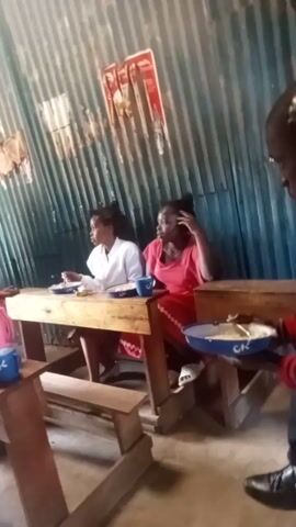 Two people are sitting at a wooden table. One is wearing a pink dress and the other a white shirt. They are inside a building with corrugated metal walls. There is a red and white advertisement on the wall behind them. Another person is standing beside the table.