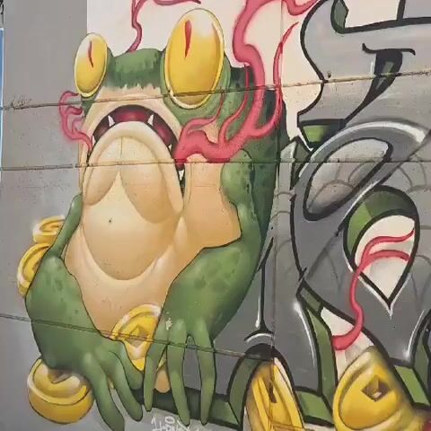 The video shows a close-up of a mural on a concrete wall. The mural features a cartoonish green frog with large yellow eyes and gold coins around it. To the right of the frog, there is a stylized, gray and green graffiti tag. Red lines or tendrils are incorporated throughout the artwork. The camera slowly pans across the mural, focusing on the different elements of the artwork. The wall is made of concrete blocks, and the graffiti appears to have been applied to the wall.