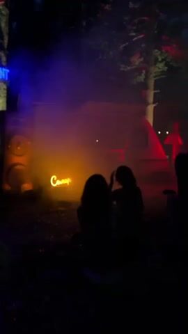 Two individuals, silhouetted from behind, are facing a neon sign that reads "Солар" which is illuminated in orange. Behind them, there is a trailer with a serving window open and illuminated with red light. To the left of the trailer is a large speaker. The scene is outdoors at night with trees visible in the background. There is smoke or fog around the trailer and neon sign, illuminated by blue and orange lights.