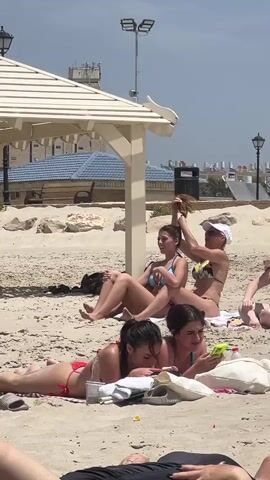 The video shows a group of women at the beach. One woman in a black and yellow bikini is sitting and pulling another woman's hair up into a ponytail. A third woman is lying down on a towel and looking at a phone. Another woman is next to her, also lying down, and looking at a phone. There is a white shade structure over them. There is a trash can behind the woman sitting up. In the distance, there are buildings with blue roofs.