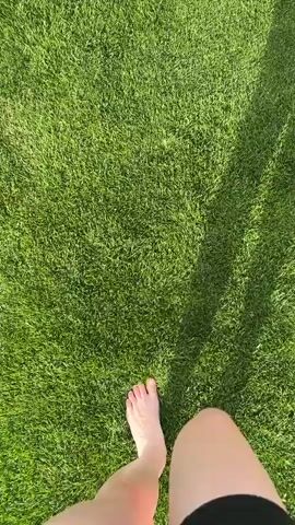 The video shows a pair of bare feet walking on bright green grass. The camera perspective is from above, looking down at the feet and the lawn. The scene is well-lit, suggesting daytime. The grass is neatly mowed and appears to be a sports field, as indicated by the presence of floodlights and a small building in the background. The feet are a person's, and they are walking away from the camera. There is a shadow on the right side of the frame, possibly from a person or structure. The video then cuts to a wider shot showing two people sitting on the grass, facing away from the camera. The man is wearing a blue t-shirt and shorts, and the woman is wearing a black top and shorts. They are on a large, manicured green field under a bright blue sky with scattered clouds. In the distance, there are hills and more sports field infrastructure. The scene is peaceful and relaxed.