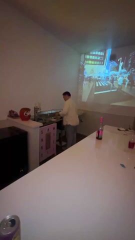 The video shows a home bar setup with a man behind it. A projector displays a busy city scene on the wall. On the white countertop, there is a can of hard seltzer and a glass. The camera pans across the bar area. There is a black tote bag with "SUPPLY LOCAL ART" printed on it. The bar has three glass-door refrigerators filled with drinks.