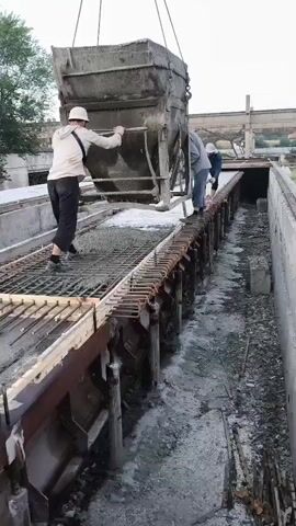The video shows a construction scene in Khasavyurt, Russia. Three men are working on a long, narrow concrete structure. A large concrete bucket is suspended from a crane, being tilted to pour concrete onto a metal grate that forms the base of the structure. One worker, wearing a white helmet and a light-colored shirt, stands on the left side of the structure, appearing to guide the concrete flow with his hands. Another worker, in a blue shirt, stands further down the structure, near the bucket, and seems to be supervising or assisting. A third worker is partially visible in the background, also near the bucket.

The setting appears to be an outdoor industrial or construction area. The concrete structure itself is long and appears to be a form for casting some type of pre-fabricated element, possibly for infrastructure like bridges or walls. The area around the structure is a mix of concrete debris, dirt, and some vegetation visible in the upper left corner of the frame. The lighting suggests it is daytime, possibly late afternoon or early morning, with soft light and no harsh shadows. The weather appears to be fair. The men are dressed in work clothes, and their focus is on the task at hand, indicating a typical workday. There is no visible text in the video.