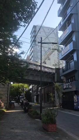 The video pans across a street in what appears to be an urban environment. In the foreground, a cobblestone sidewalk is lined with potted plants and a few parked motorcycles. A few trees with green foliage overhang the sidewalk. To the right of the sidewalk, a paved street runs parallel to it. On the right side of the street, a modern, multi-story building with blue and gray siding stands tall. A few shops are visible on the ground floor of this building. In the background, a raised train track with a train on it crosses the scene. Behind the train track, a tall, light-colored building with many windows dominates the skyline. The sky is clear and blue. The scene appears calm and ordinary, with no discernable activity or interaction between people. The overall impression is that of a typical street view in a Japanese city.