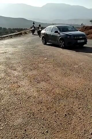 The video shows a two-lane asphalt road in a mountainous area, with two motorcycles approaching the camera. The road surface appears somewhat worn. Each motorcycle is ridden by a person wearing a helmet and protective gear. Both bikes are equipped with luggage, indicating they are likely on a tour. A car is parked on the right side of the road. Mountains are visible in the background, with a hazy sky above. The surrounding terrain is rocky and arid. The video is shot from a low angle on the side of the road, which is covered with loose gravel.