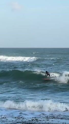 A surfer rides a wave in the ocean. The surfer is wearing dark clothing and is on a red surfboard. The water is a deep blue-green, with white foam from the waves. In the background, there is a small, rocky island with trees on it, and the sky is a pale blue with a few wispy clouds. The shore is lined with dark rocks.
