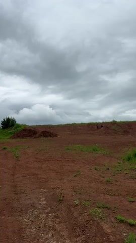 An outdoor scene features a reddish-brown dirt landscape under a cloudy sky. In the foreground, a child wearing a light-colored long-sleeved shirt with dark patterns, black pants, and a dark cap walks and then runs towards the camera, briefly raising their arms. In the mid-ground to the right, a second person in dark clothing is seated on a dirt slope. Green foliage lines the distant horizon to the left.