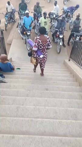 A woman walks up concrete steps while carrying a purple and white object and a tan bag. A person is seated near the stairs on the left. At the top of the stairs, a group of people is seated on motorcycles, appearing to wave and gesture. The setting appears to be outdoors.