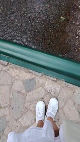 The video shows a view of a small waterfall flowing into a shallow stream. The waterfall is made of stacked stones with moss and vegetation growing on them. The stream bed is rocky and the water is clear. In the foreground, a person's feet are visible, wearing white sneakers. The person appears to be standing on a paved walkway made of stones. The scene suggests an outdoor natural setting, possibly a park or a garden, given the presence of the walkway and the landscaped waterfall. The lighting suggests it is daytime, but the weather is not explicitly indicated. There is no visible text in the video. The overall mood is peaceful and serene, focusing on the natural beauty of the waterfall and stream. The location context of Lokorsko, Bulgaria, places this scene within a specific geographical area.