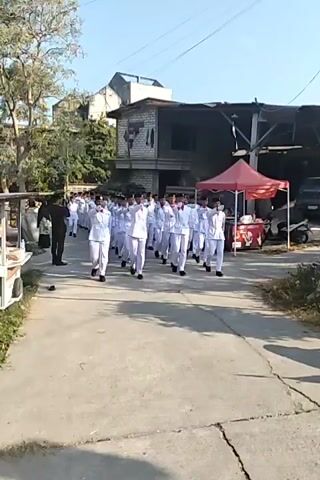 A group of young people in white uniforms with red neckerchiefs are marching in formation on a paved road under a clear sky. They are walking towards the camera, with some individuals in the front row raising their arms. In the background, there are buildings, a red tent, and parked vehicles. A few people in dark clothing are visible on the side of the road, observing the march. The scene appears to be outdoors in a somewhat industrial or less developed area, with trees and vegetation present.