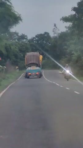 A truck carrying stacked cargo, possibly goods wrapped in corrugated material, is traveling down a two-lane paved road surrounded by lush green trees. The truck is in the right lane, with another vehicle, possibly a car, partially visible on the left side of the road near the truck. A motorcycle with its headlight on is approaching the truck from the opposite direction, in the left lane. The sky is overcast, suggesting a cloudy or possibly rainy day. The road has white dashed lines marking the lanes. The video appears to be shot from a moving vehicle.