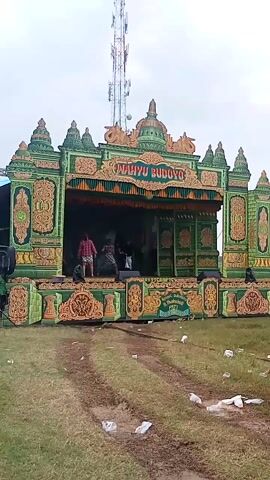 Here is a concise description of the video:

The video captures a stage adorned with intricate green and gold decorations, with the words "Wahyue Budoyo" displayed prominently. Inside, several people are visible, possibly setting up or preparing for a performance. The stage is set on a grassy area with some litter strewn around. In the background, a tall telecommunications tower stands. The video is taken from an upward, slightly angled perspective.