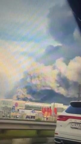 This media file depicts a large plume of smoke rising into the sky, suggesting a significant fire or industrial incident. The scene appears to be captured from a moving vehicle, possibly on a highway, given the presence of a guardrail in the foreground and other vehicles.

The subjects include a white SUV in the foreground, with a license plate visible: K973EA. Behind it, a truck with a trailer is partially obscured by the smoke. In the middle ground, several low-rise buildings are visible, some with red accents. The overall setting seems to be an urban or industrial area, with the smoke dominating the background.

The activity is the ongoing plume of smoke, which is dense and billowing, suggesting a large-scale event. The time of day appears to be daytime, judging by the bright sky, though the smoke creates a somewhat overcast or hazy atmosphere. The weather is not clearly discernible due to the smoke, but the sky is a mixture of blue and grey, with hints of sunlight.

Notable details include the scale of the smoke, which suggests a serious incident. The presence of multiple vehicles implies that this is a public area, likely accessible by road. No specific emotions are conveyed, but the image of a large smoke plume can evoke concern or alarm.

There is some visible text on the license plate of the white SUV, reading "K973EA". The location context provided is Sof'ino, Russia.