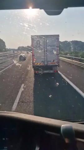 The video shows a view from the driver's seat of a vehicle on a highway. The sun is bright and reflects off the windshield, which has a lot of streaks and smudges on it. In front of the vehicle, there is a large white semi-trailer truck with text on the back. To the left of the truck, there are other vehicles on the highway. The highway has multiple lanes and a concrete barrier on the left. The sun is high in the sky, suggesting it is daytime. The video appears to be a first-person perspective of driving on a highway.